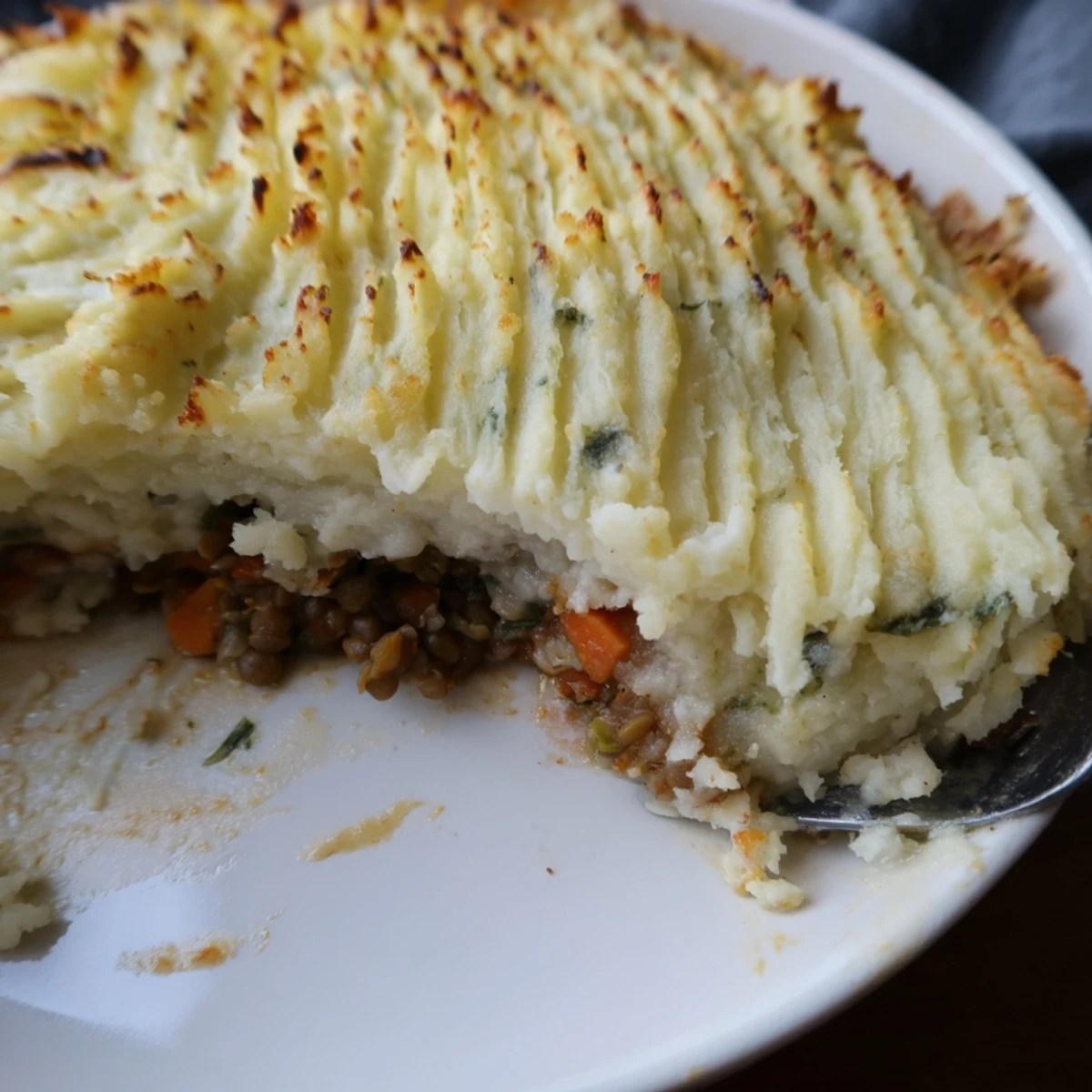 A slice of Vegan Lentil Shepherds Pie with Fluffy Mash is lifted from the baking dish, revealing layers of savory lentil and vegetable filling beneath golden, fork-crested potatoes.
