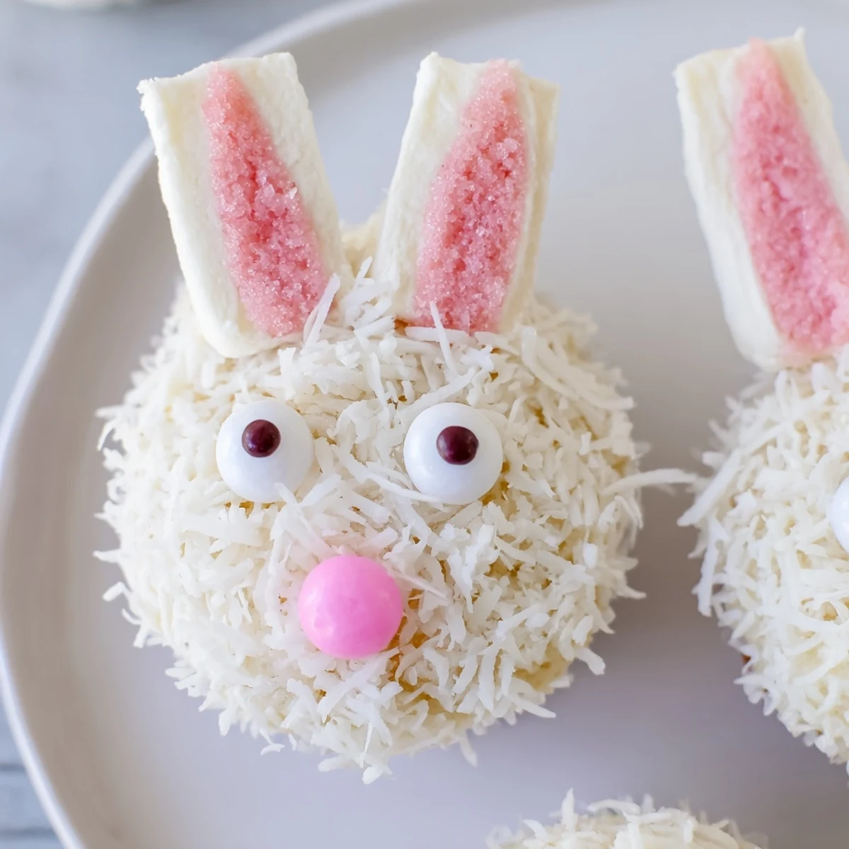Spring-themed vanilla bunny cupcakes featuring candy eyes, pink sugar-dipped ears, and coconut coating