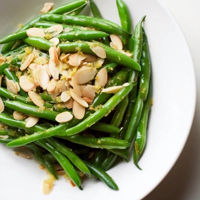 Tender green beans with lemon and almonds in a skillet, with garlic butter and fresh lemon zest nearby.